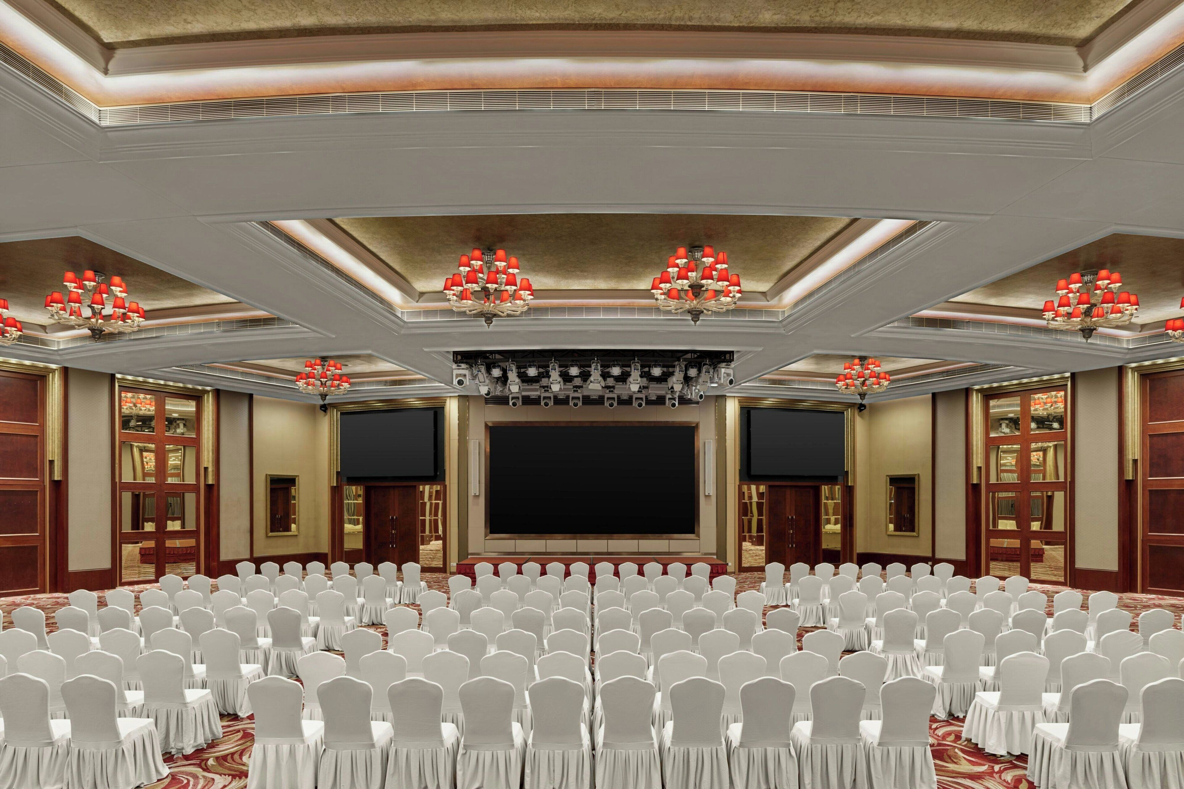 A large conference room with rows of white chairs, a stage with a black screen, and red chandeliers.