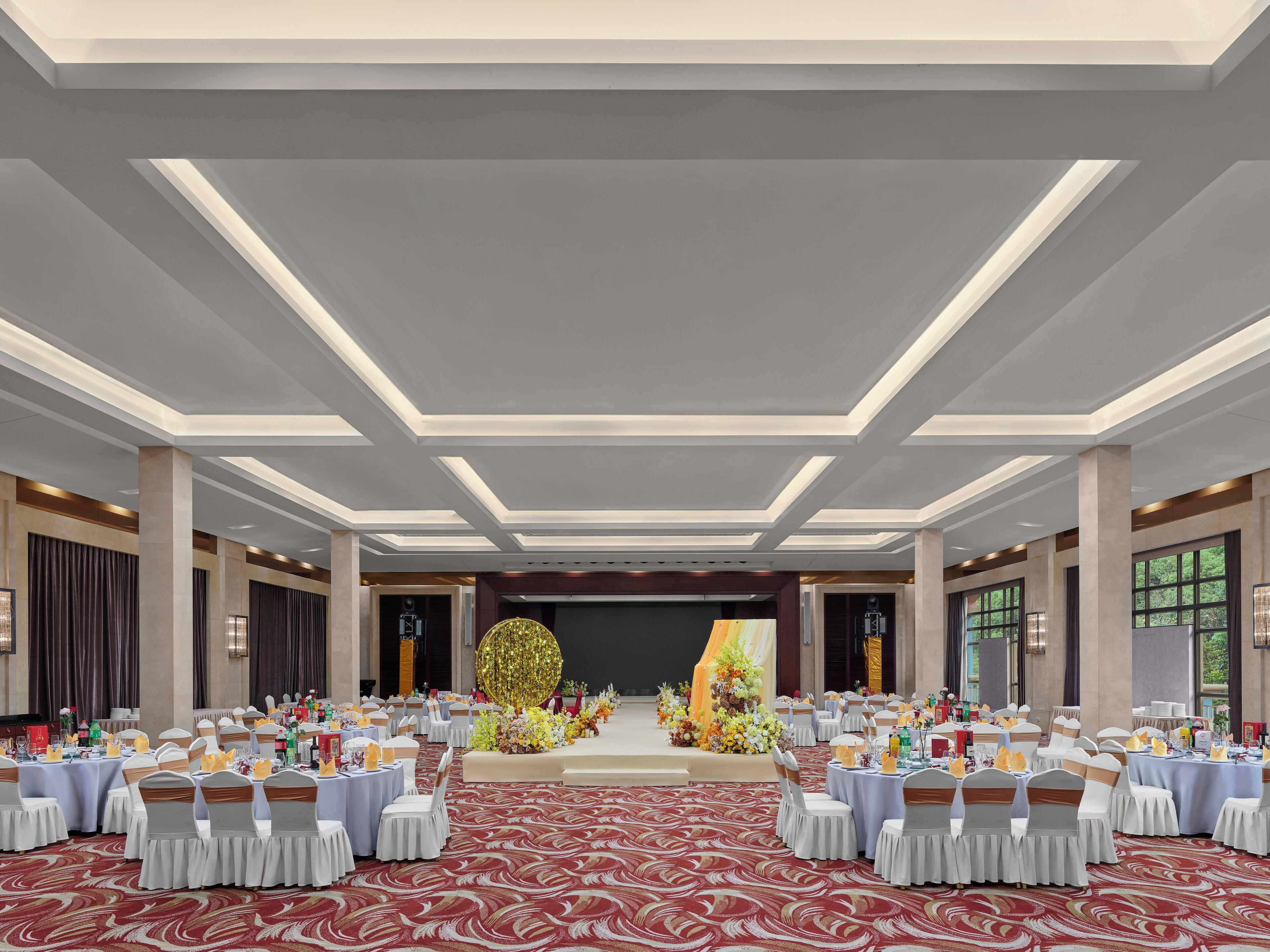 A banquet hall with round tables, white chairs, and a stage with floral decorations.