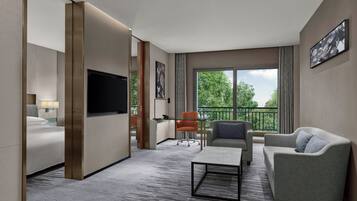 Habitación de hotel con cama, sofá, mesa de centro, televisor, escritorio y una silla. Tiene un balcón con vistas a árboles.