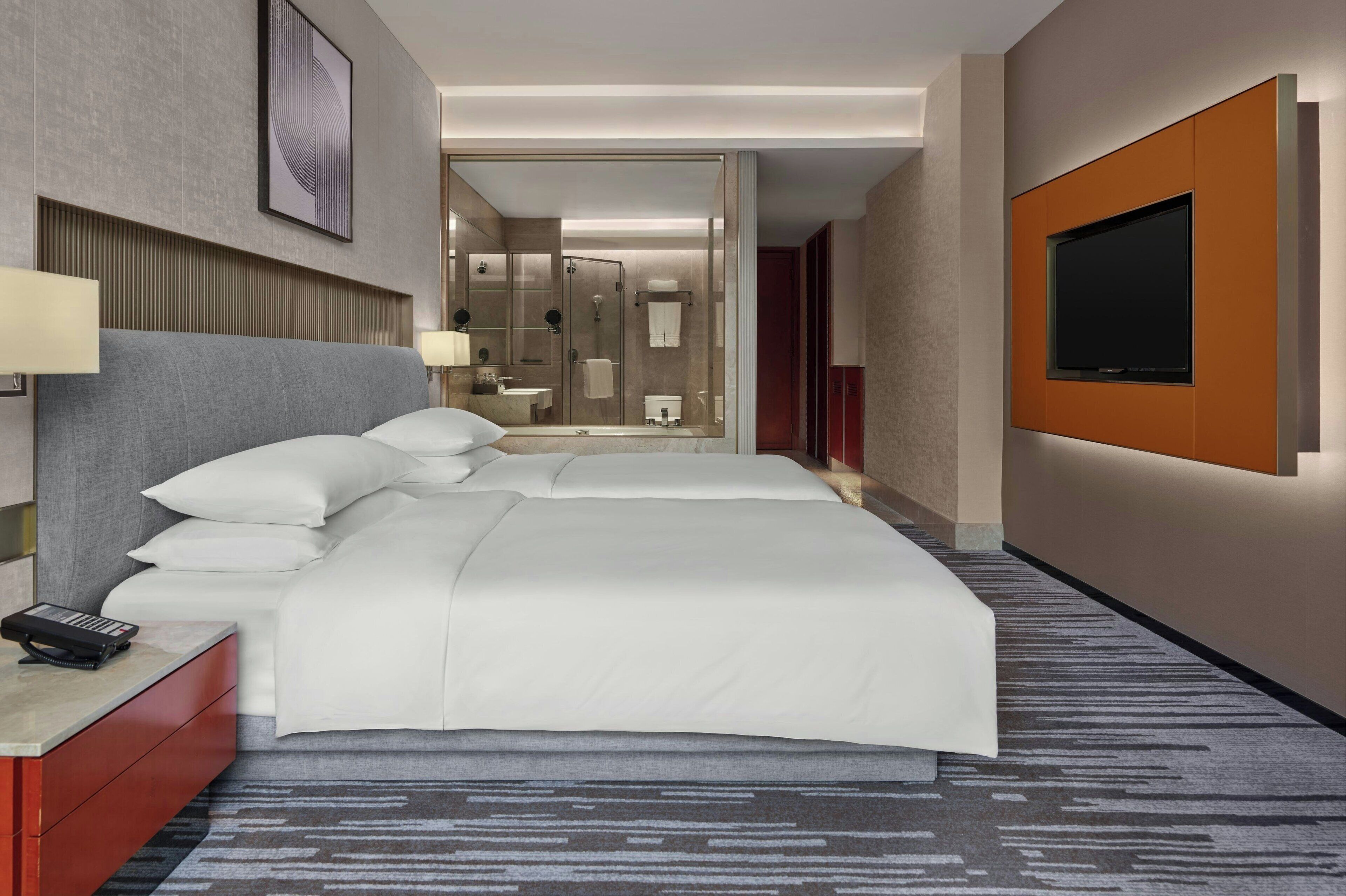 A hotel room with a large bed, a TV mounted on the wall, a bedside table with a lamp, and a bathroom visible through an open door.