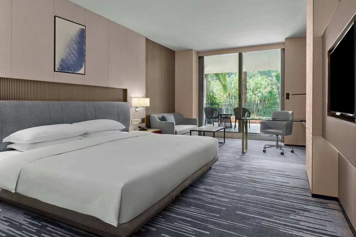 Ein Hotelzimmer mit einem großen Bett, einem Schreibtisch mit Stuhl, einem Fernseher und einem Balkon mit Blick auf Palmen.