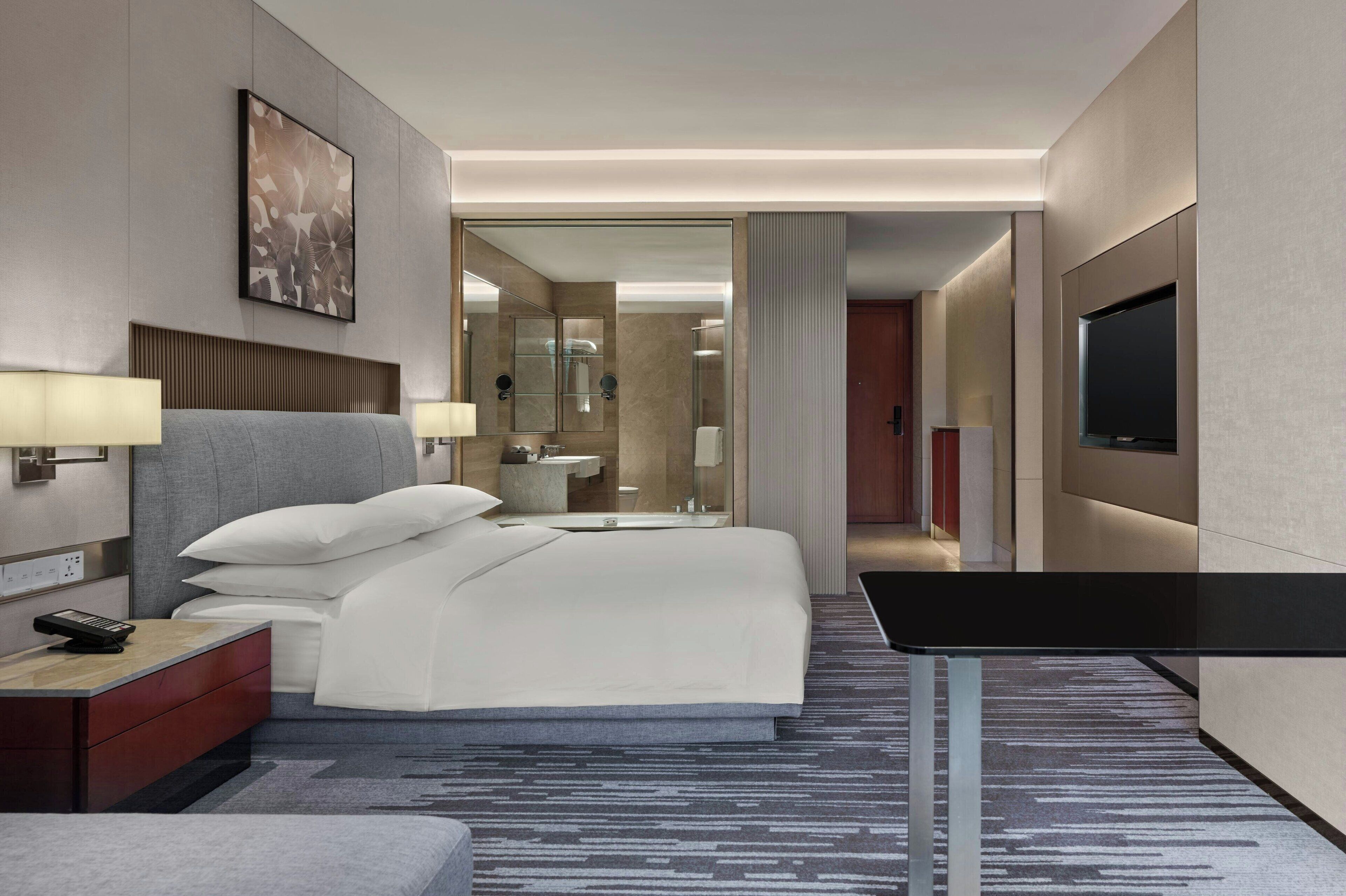 A modern hotel room with a large bed, bedside lamps, a flat-screen TV, a desk, and a bathroom visible through an open door.