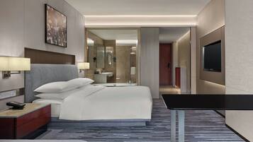 A modern hotel room with a large bed, bedside lamps, a flat-screen TV, a desk, and a bathroom visible through an open door.