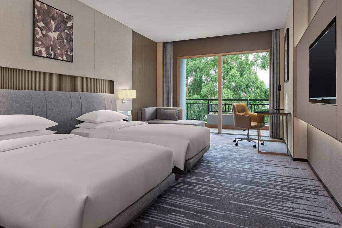 Ein Hotelzimmer mit zwei grossen Betten, einem Schreibtisch mit Stuhl, einem Fernseher und einem Balkon mit Blick auf Bäume.