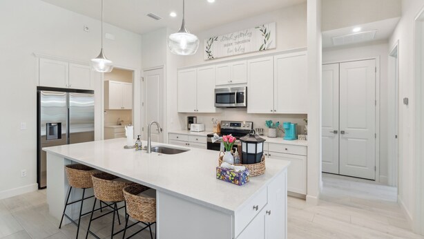 Una cocina moderna con armarios blancos, una isla central con taburetes, electrodomésticos de acero inoxidable y un letrero decorativo en la pared.