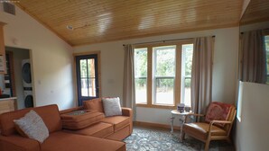 Una sala de estar acogedora con un sofá modular, una mesita y un sillón. Amplias ventanas con cortinas, un tapete estampado y un techo de madera.