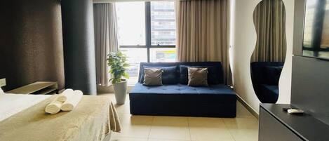 Habitación de hotel con cama, sofá, televisión, planta en maceta y ventana con cortinas.