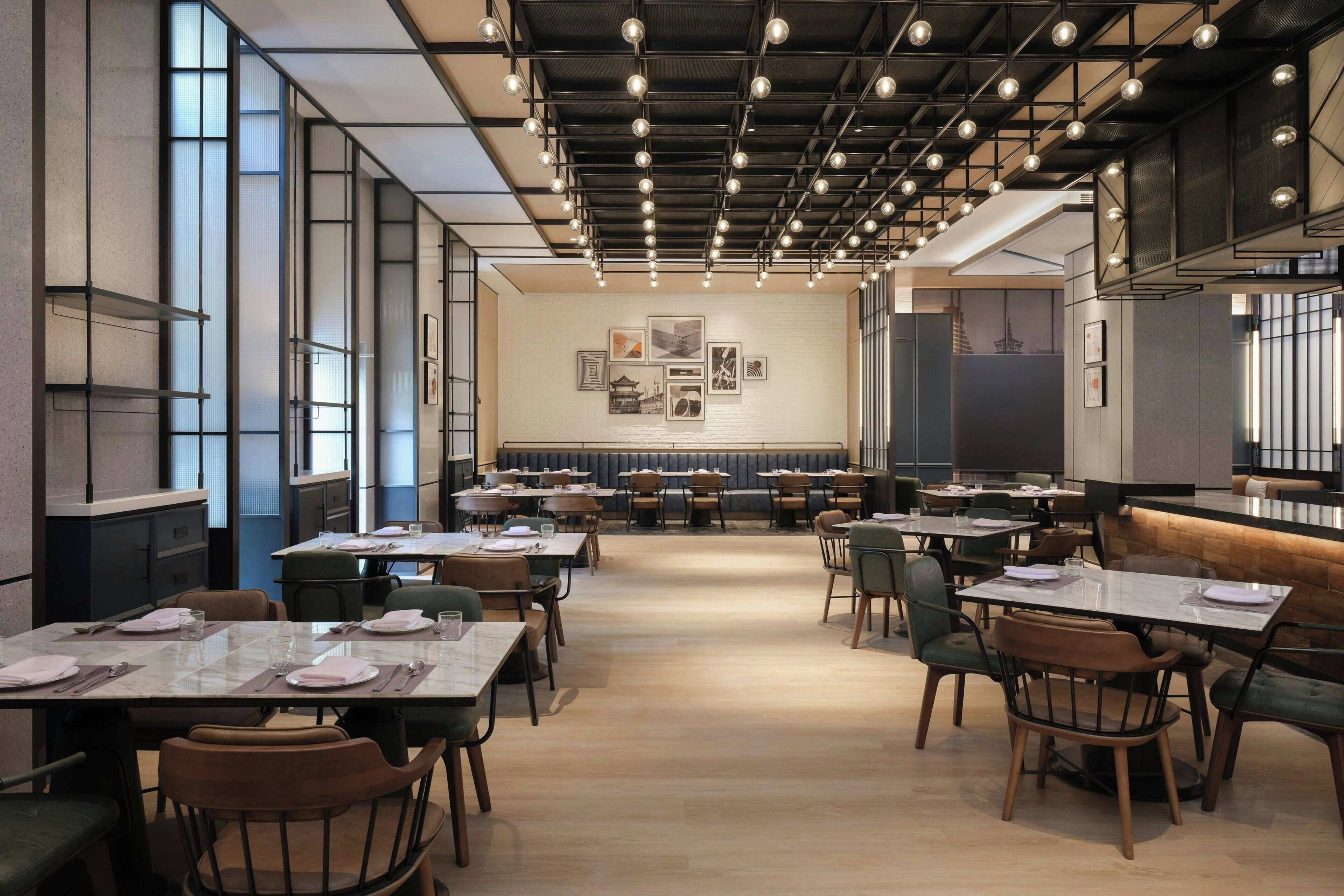 Un espace de restauration moderne, doté de plusieurs tables et chaises, d’un plafond haut avec des poutres apparentes et des luminaires, ainsi que d’un mur orné de tableaux encadrés.