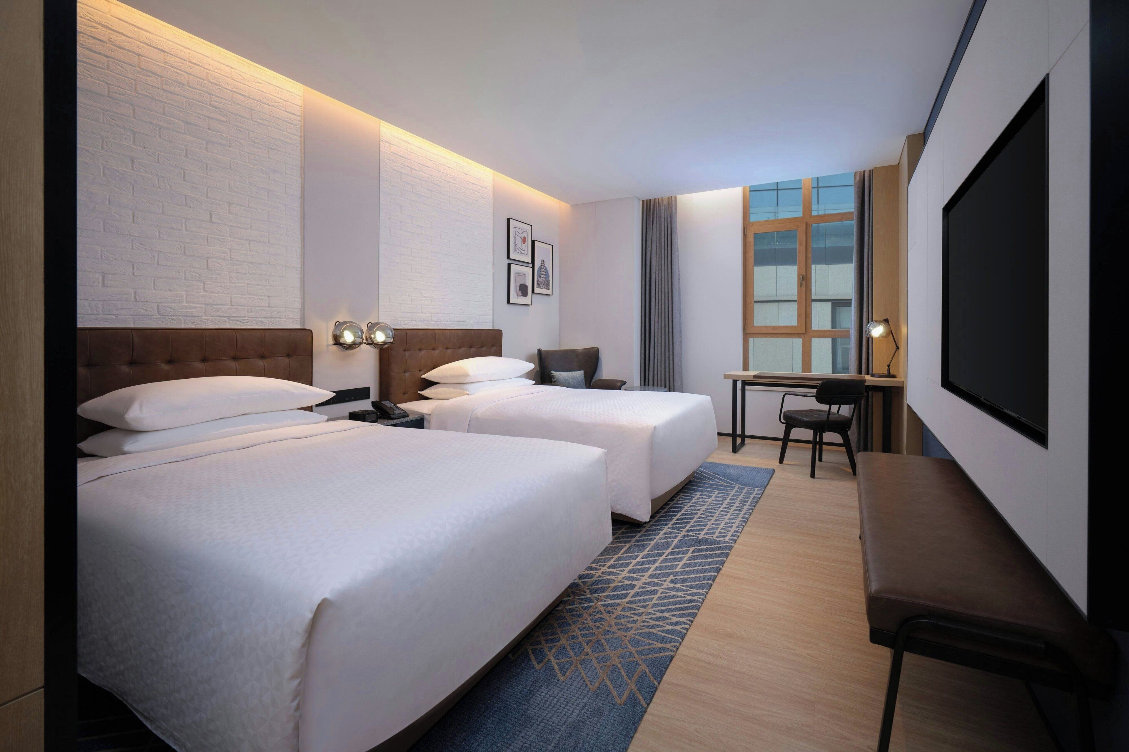 A hotel room with two beds, a desk, a chair, a TV, and a window with curtains.