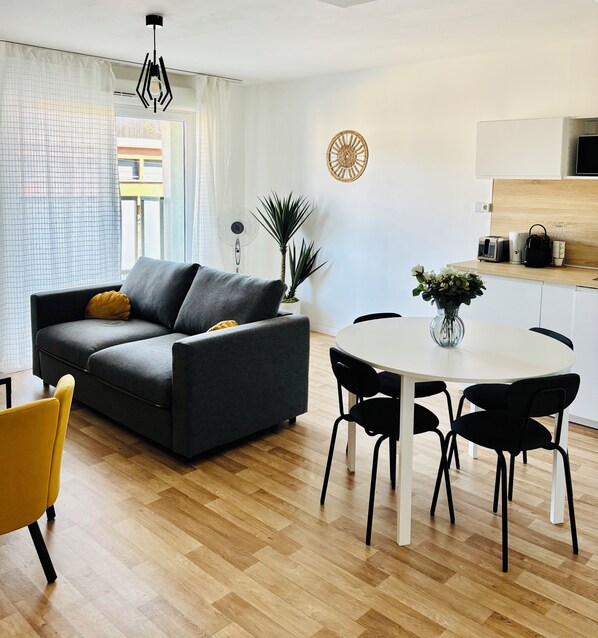 Una sala de estar moderna con un sofá gris, una butaca amarilla, una mesa de comedor con sillas negras, un ventilador de techo blanco y una zona de cocina con microondas y tostadora.
