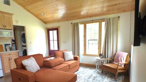 Un salón acogedor con techo de madera, un sofá, una silla, una mesita y una ventana con cortinas.