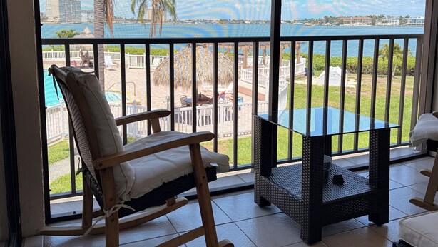 Un balcon avec un fauteuil à bascule, une table et une vue sur une piscine et des palmiers.