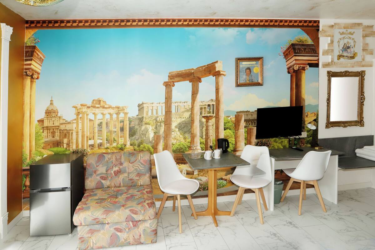 A room with a mural of ancient ruins, a sofa, a dining table with chairs, a TV, and a mirror.