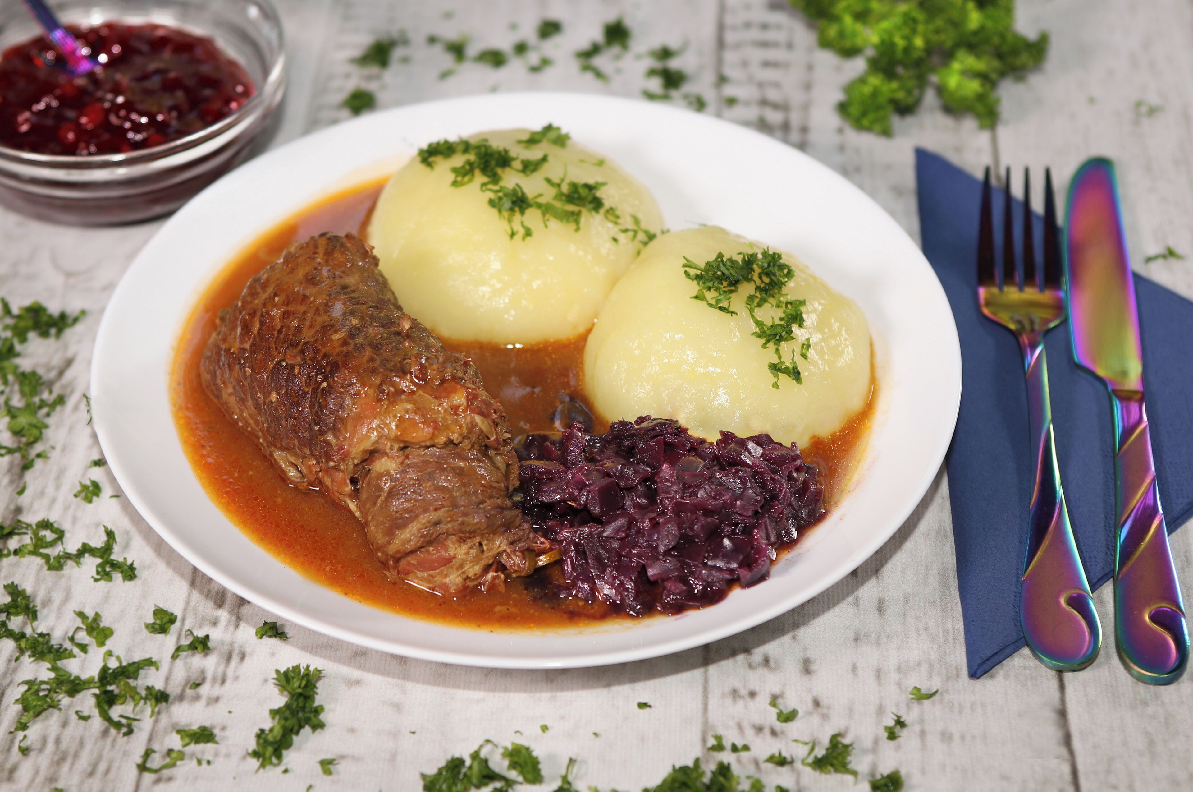 A plate with meat, potatoes, and red cabbage, accompanied by a fork and knife on a napkin.