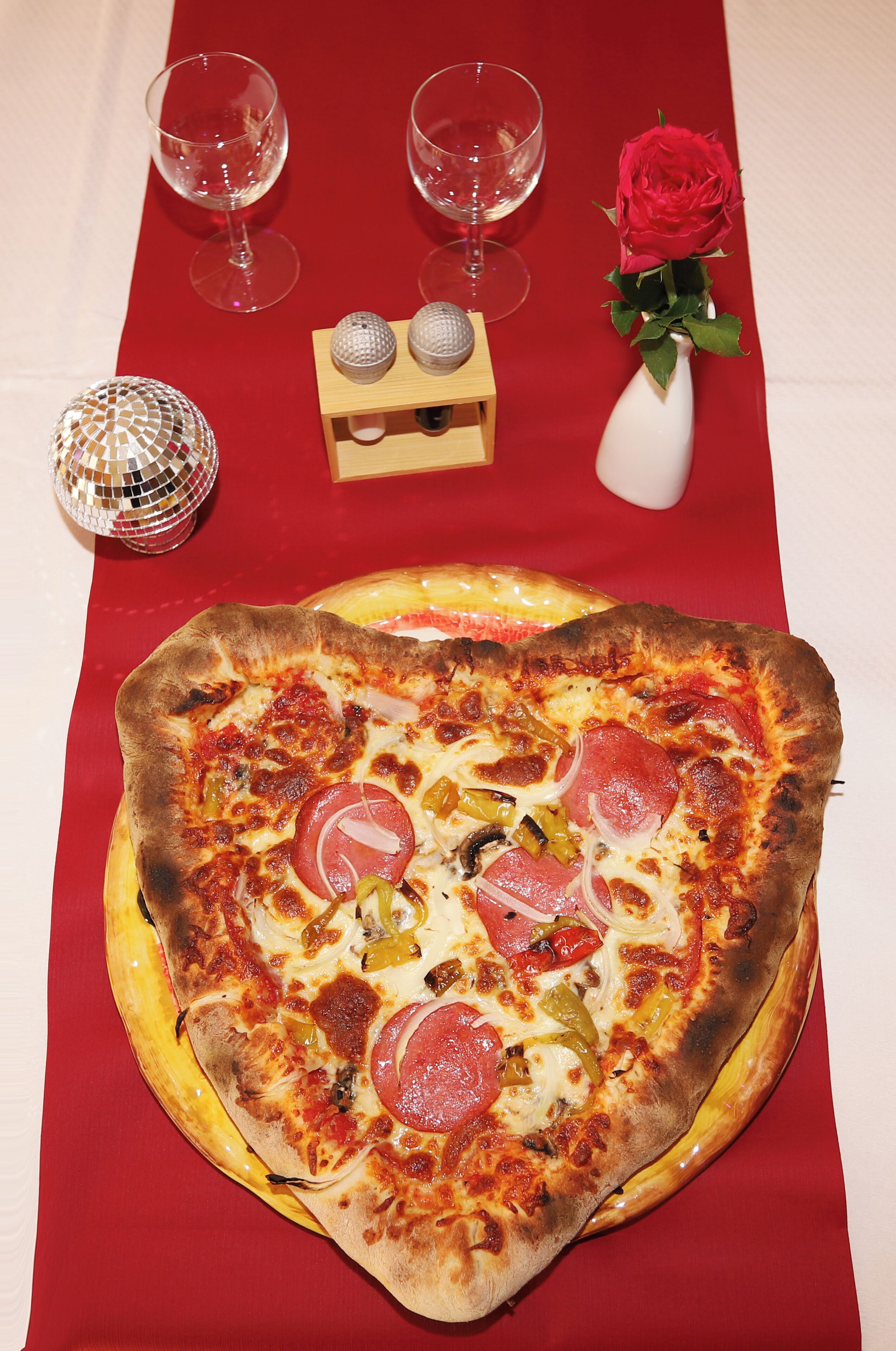 Une pizza en forme de cœur posée sur une nappe rouge, entourée de verres à vin, d’une rose et de deux boules décoratives.