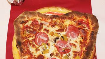 Une pizza en forme de cœur posée sur une nappe rouge, entourée de verres à vin, d’une rose et de deux boules décoratives.