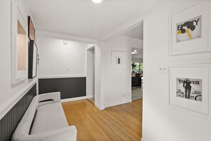 Un pasillo con piso de madera, paredes blancas y un banco blanco. Hay cuadros enmarcados en las paredes y una puerta que da a otra habitación.