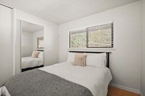 A bedroom with a bed, a window with blinds, a mirror, and a wooden floor.