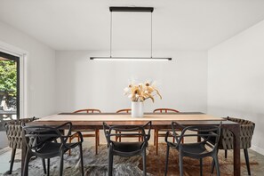 Un comedor moderno con una mesa de madera, seis sillas, una lámpara colgante y un jarrón con flores secas.