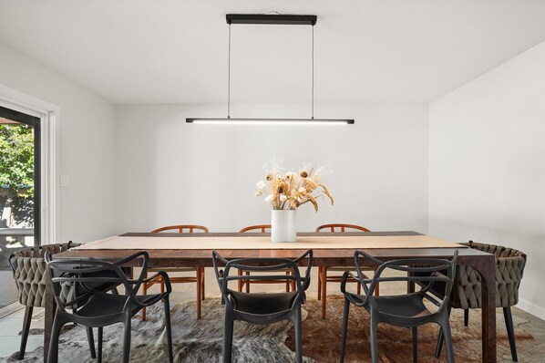 A modern dining room with a wooden table, six chairs, a hanging light fixture, and a vase with dried flowers.
