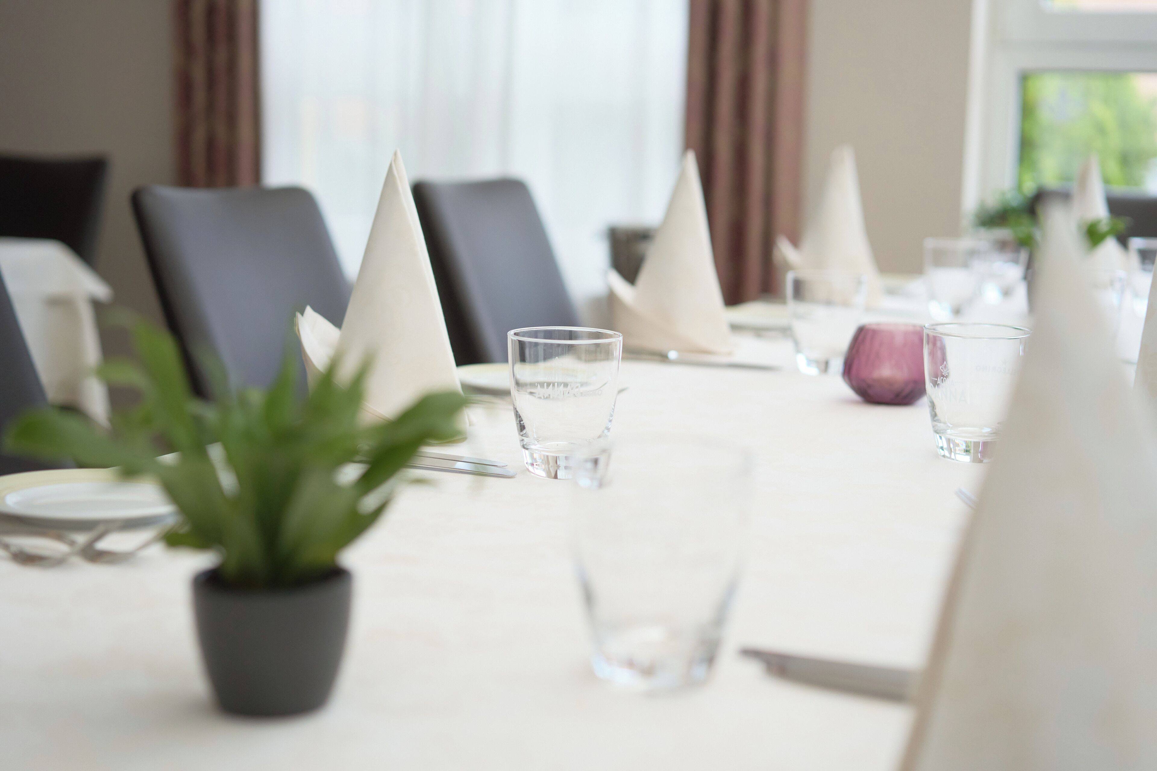 A dining table set with white napkins, clear glasses, and a potted plant.