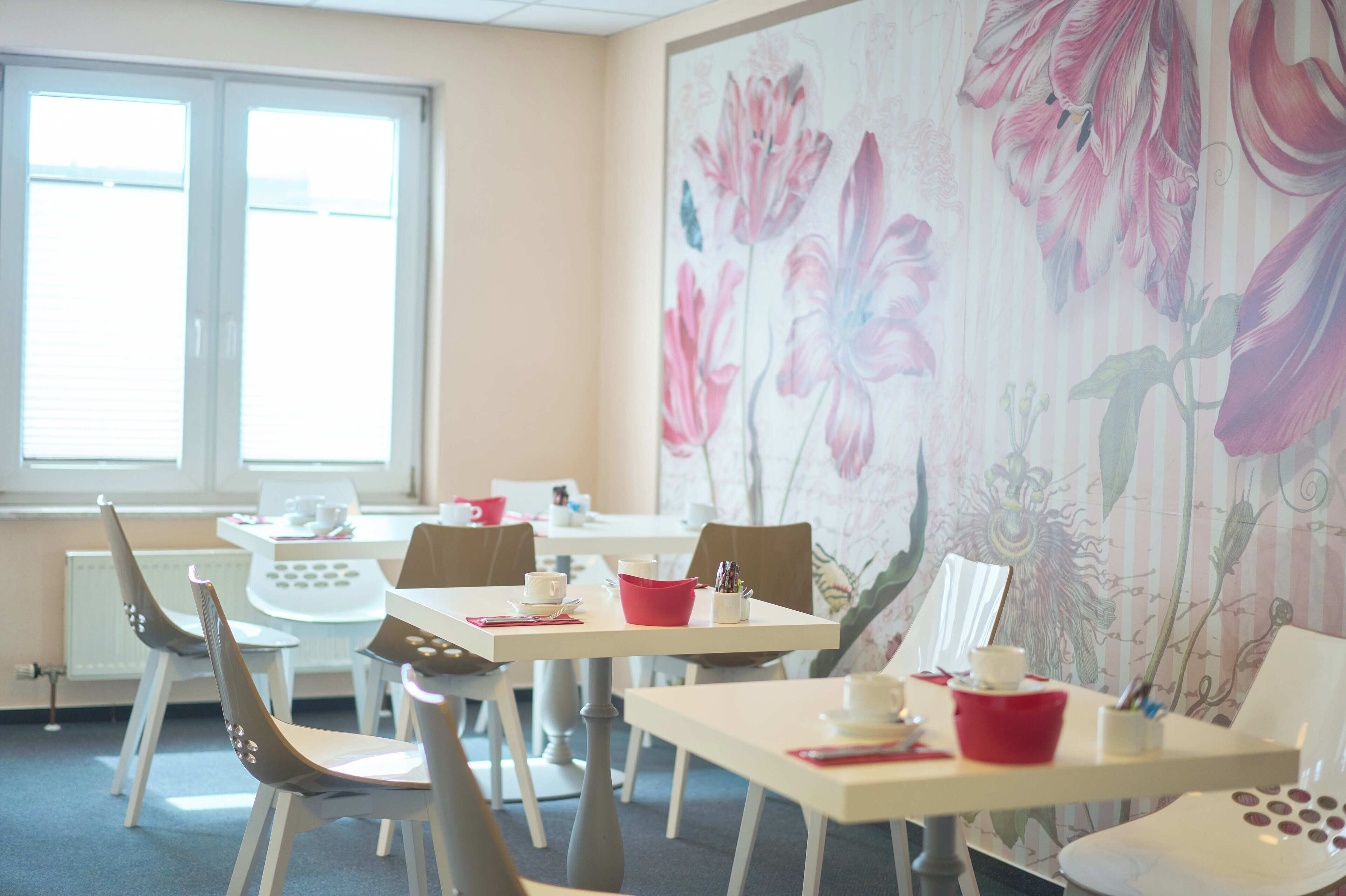 Een eethoek met witte tafels en stoelen, bloemen behang en een raam met jaloezieën.