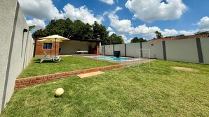 A backyard with a swimming pool, a patio with chairs, and a brick wall.