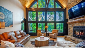 A spacious living room with a large stone fireplace, a leather sofa, and a large window overlooking a forest.