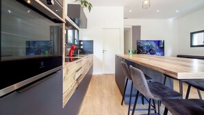 A modern kitchen with a built-in oven, a large TV, and bar stools. - Maison Marguerite - Welkeys (Bayonne)