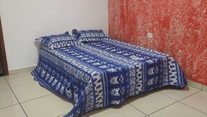 Un petit lit avec une couvre-lit bleue à motifs dans une pièce dont un mur est rouge et présente une texture particulière, et dont le sol est carrelé.