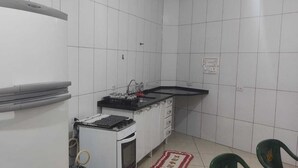 Une cuisine compacte avec des murs recouverts de carreaux blancs, un réfrigérateur blanc, une petite cuisinière et un évier doté d’un plan de travail noir.