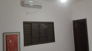 Une pièce avec un mur blanc, une fenêtre avec des stores horizontaux, une porte rouge et un climatiseur fixé au mur.