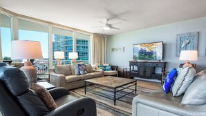 A modern living room with a sofa, armchairs, a coffee table, and a TV. There are large windows with a view of a building and a ceiling fan. - Four Bedroom Condo with Panoramic Views and Large Wraparound Balcony. Complimentary Boat Slip! (Orange Beach)