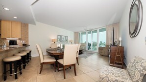 A modern living room with a dining area, a kitchen with bar stools, and a large window offering a view of the outdoors. - Cozy Three Bedroom Poolside Condo. Complimentary Boat Slip! (Orange Beach)