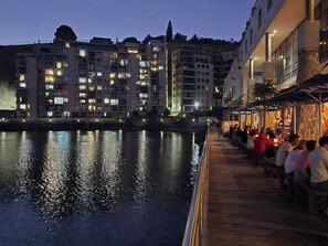 Un paseo marítimo con una pasarela de madera, personas sentadas en mesas y edificios con ventanas iluminadas al fondo.