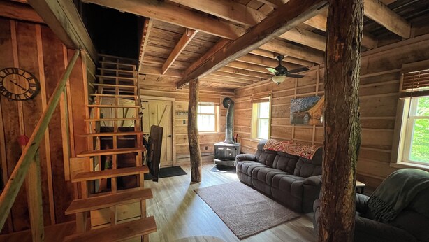L’intérieur d’une cabane en rondins, avec un escalier en bois, un canapé, une cheminée et une horloge au mur.