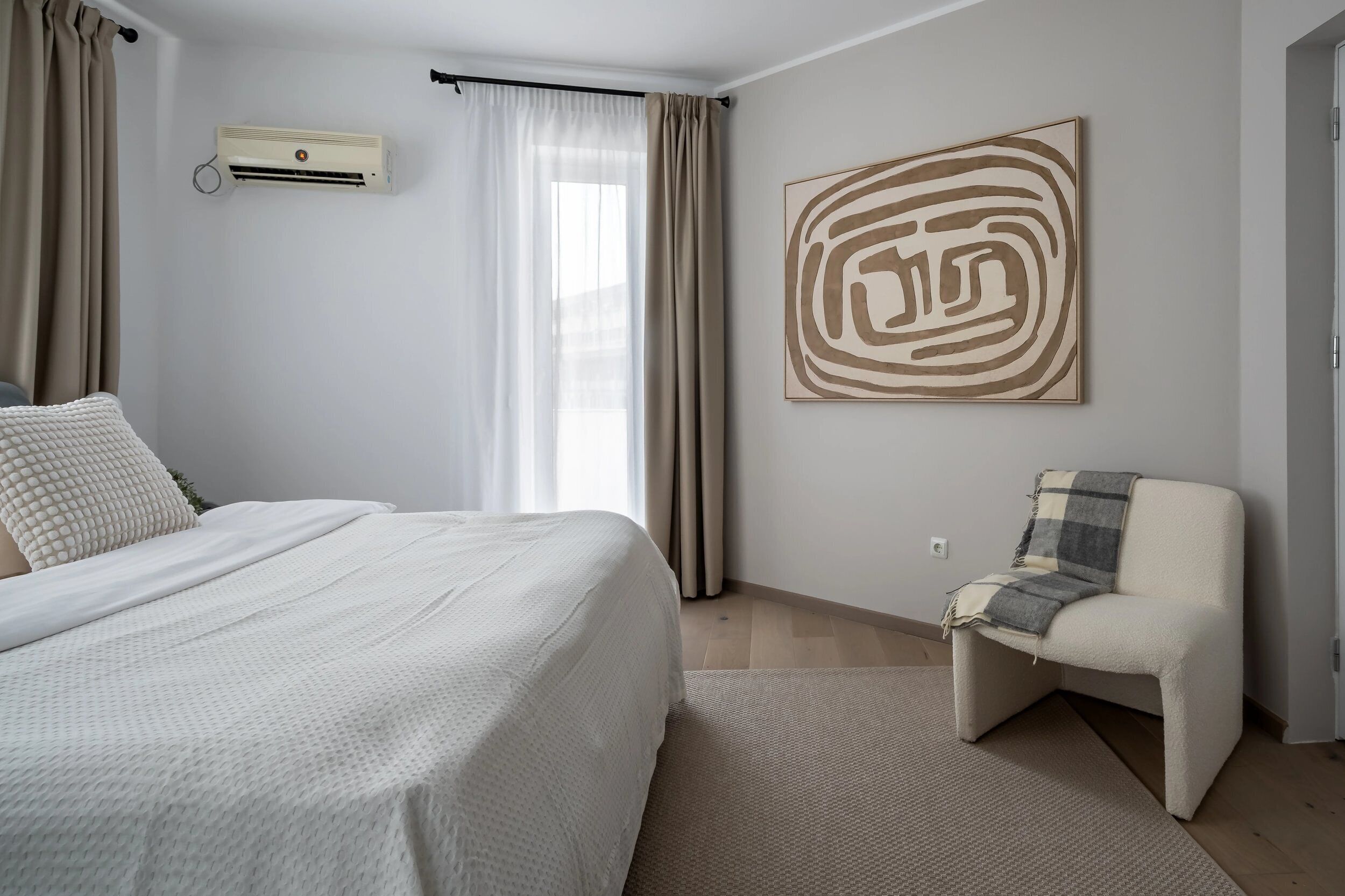A bedroom with a bed, a chair, a wall-mounted air conditioner, and a large abstract painting.