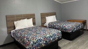 A room with two beds, each with a wooden headboard and a floral bedspread. There is a small wooden table beside the beds.