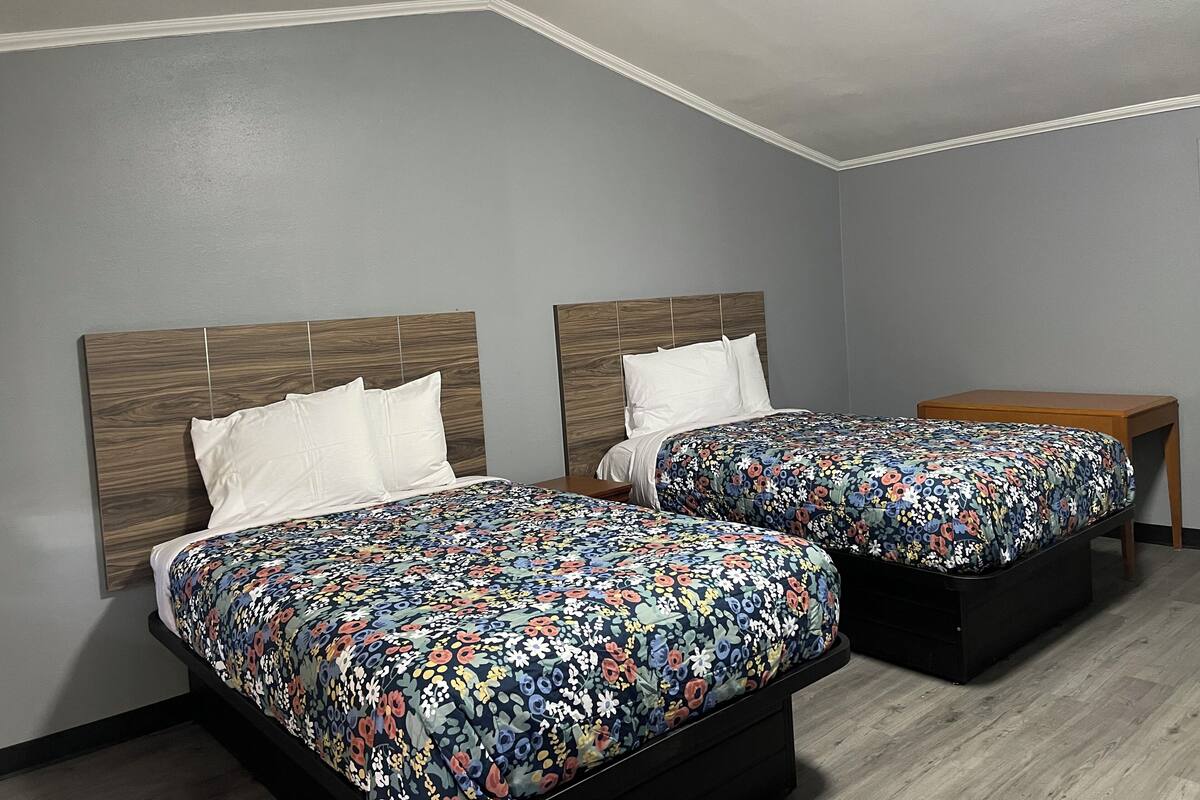 A room with two beds, each with a wooden headboard and a floral bedspread. There is a small wooden table beside the beds.