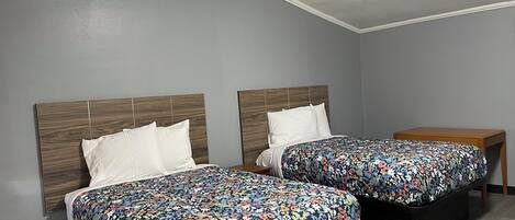 A room with two beds, each with a wooden headboard and a floral bedspread. There is a small wooden table beside the beds.