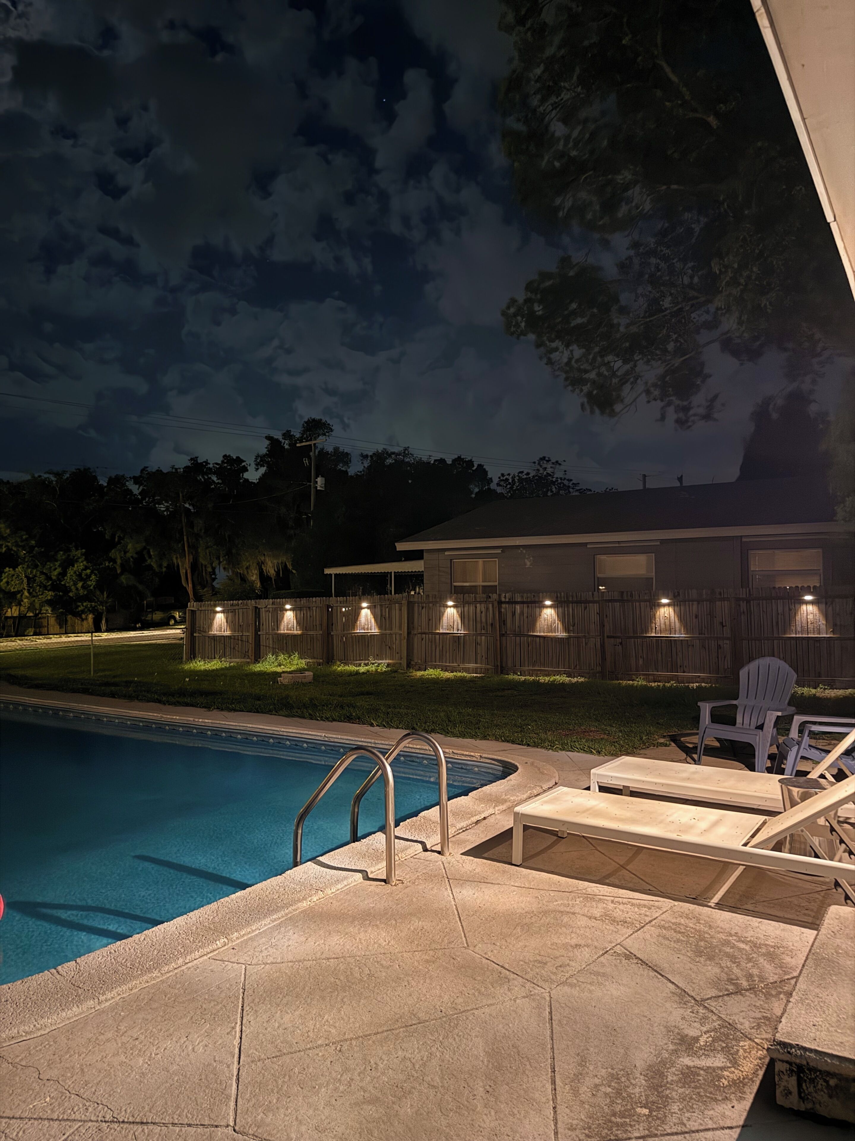 A swimming pool with a metal ladder, lounge chairs, and a wooden fence with lights.