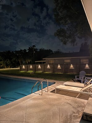 A swimming pool with a metal ladder, lounge chairs, and a wooden fence with lights.