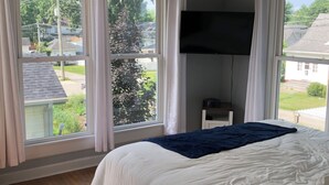 A bedroom with a bed, a ceiling fan, a TV mounted on the wall, and large windows with curtains.