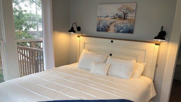 A neatly made bed with white linens and a blue throw blanket, two bedside lamps, and a painting of a rural scene above the bed.