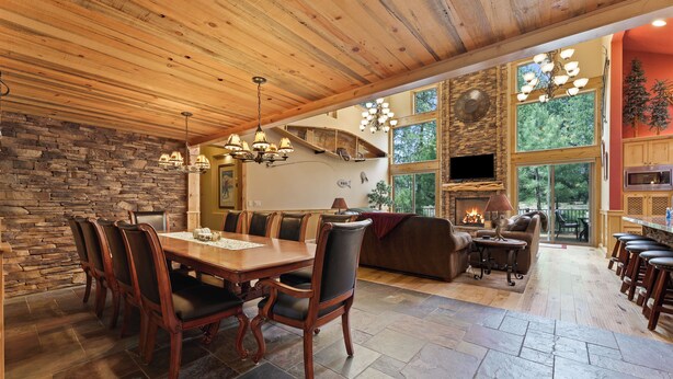 A spacious dining area with a wooden table and chairs, a stone wall, and a large window overlooking a forest. - 4 Red Fir - Ultimate Sunriver Stay, Game Room, Hot Tub, Walk to SHARC, Passes Included (Sunriver)