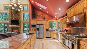 A spacious kitchen with wooden cabinets, a large island, stainless steel appliances, and a stone fireplace. - 4 Red Fir - Ultimate Sunriver Stay, Game Room, Hot Tub, Walk to SHARC, Passes Included (Sunriver)
