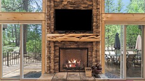 A stone fireplace with a flat-screen TV above it, surrounded by large glass doors leading to an outdoor deck. - 4 Red Fir - Ultimate Sunriver Stay, Game Room, Hot Tub, Walk to SHARC, Passes Included (Sunriver)
