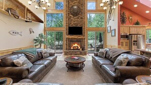 A spacious living room with leather sofas, a stone fireplace, and large windows offering a view of trees. - 4 Red Fir - Ultimate Sunriver Stay, Game Room, Hot Tub, Walk to SHARC, Passes Included (Sunriver)