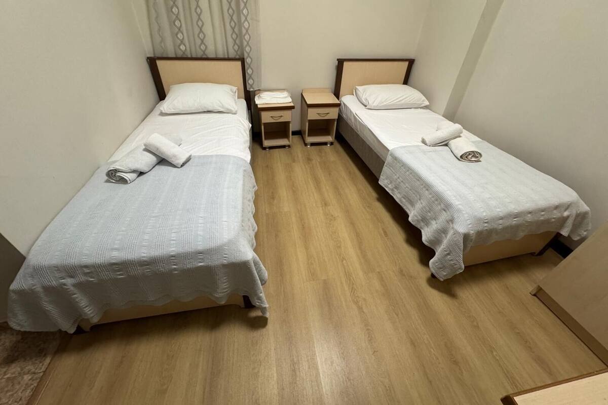 A hotel room with two single beds, each with a white bedspread and a towel. There is a wooden nightstand between the beds, and a small drawer unit to the right.