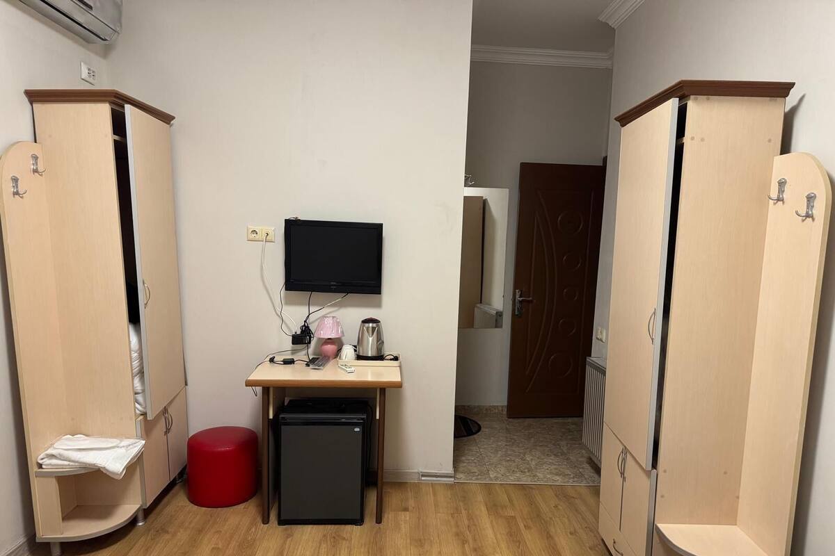 A compact room with two wooden wardrobes, a small desk with a TV, a mini fridge, and a red stool.